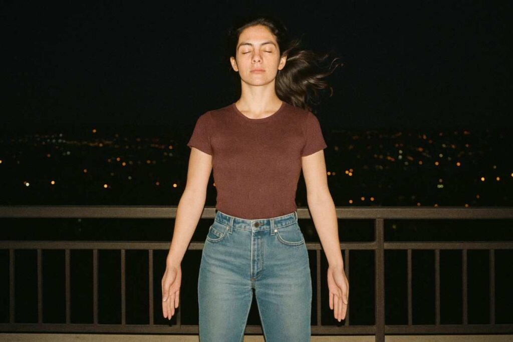 Balcony night flash photo from the 1990s, a person standing confidently in mountain pose with city lights behind, wind in hair, faded tones, spiritual and bold, grainy camera texture, low resolution but emotive image, feeling of transformation

