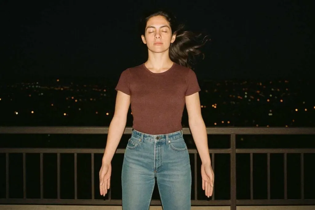 Balcony night flash photo from the 1990s, a person standing confidently in mountain pose with city lights behind, wind in hair, faded tones, spiritual and bold, grainy camera texture, low resolution but emotive image, feeling of transformation
